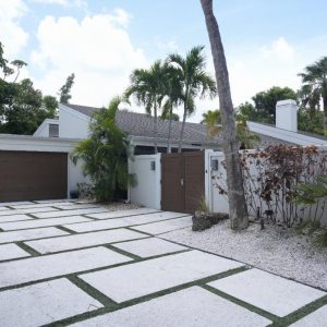 Modern white house with brown garage doors, surrounded by palm trees and a patterned driveway with green grass strips.