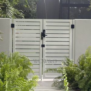 White slatted metal gate set in a stucco wall, surrounded by green ferns and tropical plants.