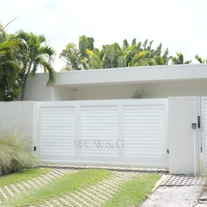 Modern white gate and driveway entrance to a house surrounded by tropical plants and greenery.