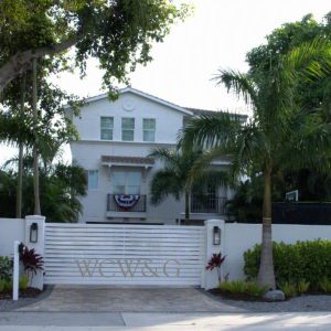 White two-story house with palm trees, gated entryway, and a patriotic banner hanging above the front door.