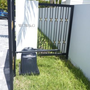Black metal gate with automatic opener, white post labeled “WCW&G,” green grass, palm trees in background.