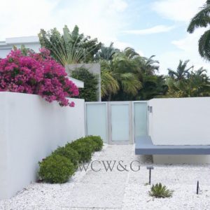 Modern white courtyard with a floating bench, gravel ground, pink bougainvillea, and tropical plants in the background.