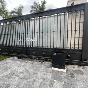 Black iron gate with “WCW&C” letters, gray stone wall behind, and a keypad panel in front on a stone driveway.