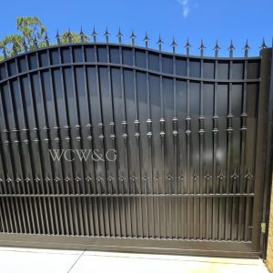 Black wrought iron gate with pointed tips, set between yellow walls, under a sunny blue sky. Letters “WCW&G” on gate.