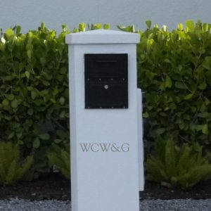 A white mailbox pillar with the letters “WCW&G” in gold, set in front of green shrubs and a white wall.