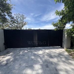 Black metal gate with solid panels and columns, set between trees under a blue sky, viewed from a paved driveway.