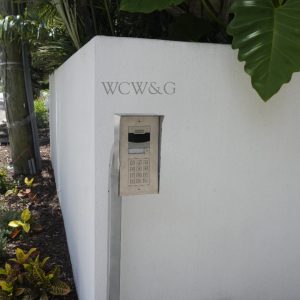 A keypad entry system is mounted on a white wall labeled “WCW&G” beside a landscaped sidewalk.
