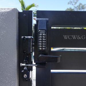 Black metal gate with a digital combination lock and “WCW&G” engraved on the gate, next to a concrete wall.