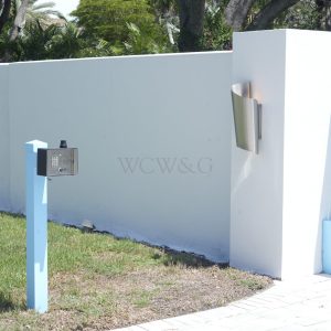 Blue gate with electronic keypad entry system on a post and a modern wall lamp next to a white wall.