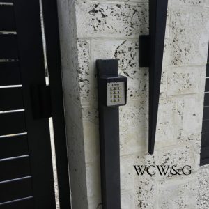A keypad entry system is mounted on a post next to a stone wall and black metal gate.