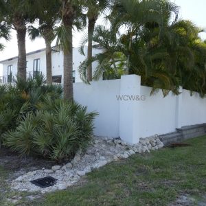 White wall with WCW&G text, surrounded by palm trees and shrubs, adjacent to a modern white building.