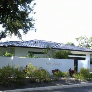 Modern single-story house with a metal roof, white wall, gate, and landscaping, viewed from the street.