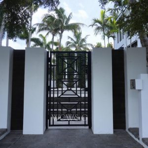 Black metal gate set in a white wall, with palm trees and a modern building visible in the background.
