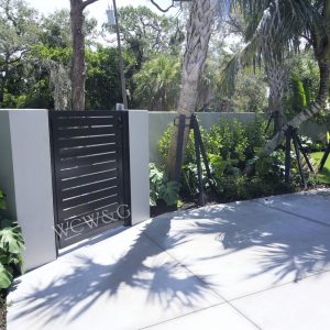 Black metal gate set in a white wall, surrounded by lush greenery and trees, with shadows on a concrete path.