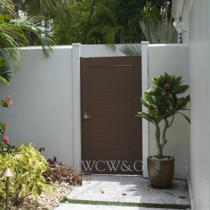 Brown wooden gate set in a white wall, next to a potted plant and surrounded by tropical greenery.