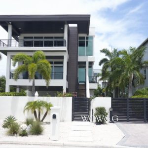 Modern three-story house with large windows, palm trees, and a black gate under a partly cloudy sky.