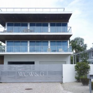 Modern three-story glass house with balconies, palm trees, and a gated entrance labeled “WCW&CO” in front.