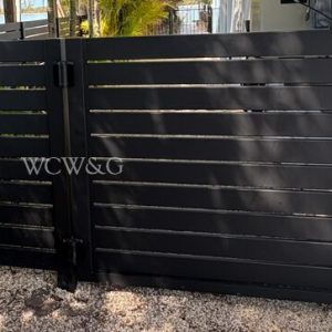 Black horizontal slat metal gate with latch, set on gravel, next to a white wall and some outdoor plants.