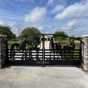 Black metal gate with animal silhouettes, flanked by stone pillars, leading to a house and driveway under a cloudy sky.