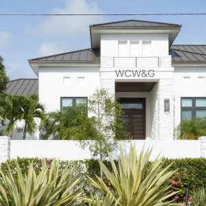 Modern white house with dark roof, tropical landscaping, and “WCW&G” sign above the front entrance.