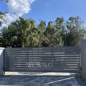 Gray metal gate with “WCW&G” letters, surrounded by trees, under a blue sky with scattered clouds.