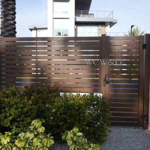 Modern brown slatted gate with “WCW&G” sign in front of a contemporary building, surrounded by greenery.