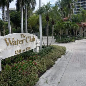 Entrance to The Water Club, featuring a white sign, palm trees, and a paved driveway leading to a gated area.