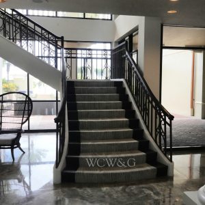 Grand staircase with black railings, striped carpet, marble floor, and large windows in a bright lobby.