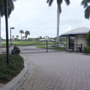 Gated entrance with a security booth and palm trees, leading to a property or community with a paved driveway.