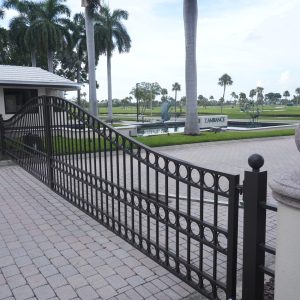Black metal gate at the entrance of a landscaped property with palm trees and a security booth.